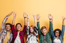 Six young people in a row of various ethnicities, laughing with arms in the air, against a yellow background.