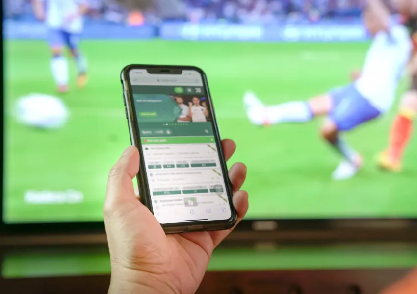 Someone holding a mobile phone, on which a betting app is open, with football on screen in the background.