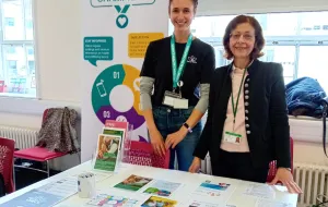 Two women smiling at a table with health leaflets. Sign reads Barnet Health Champions. 