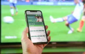 Someone holding a mobile phone, on which a betting app is open, with football on screen in the background.